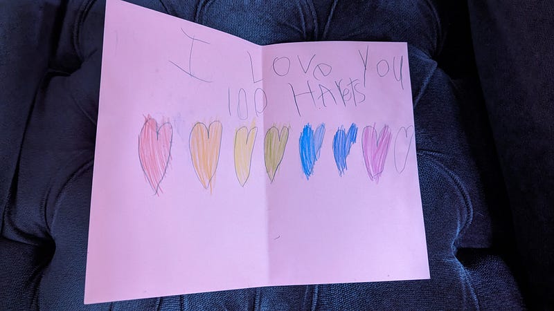 Pink paper with children’s writing I love you 100 harets followed by a line of drawn hearts in the colors of the rainbow.