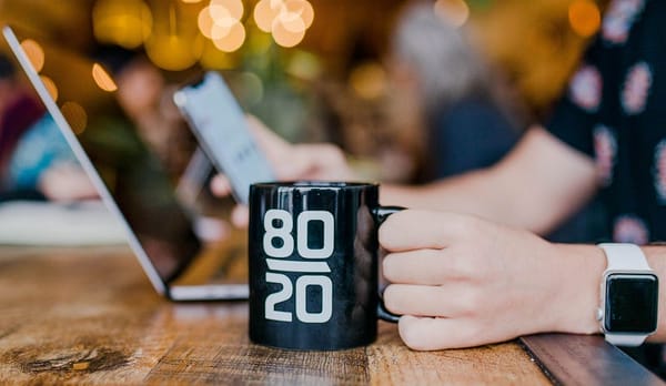 A hand grasps a mug displaying 80 20 written on its side in the foreground. In the background the human is looking at their p