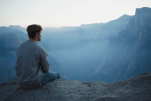 A man wearing comfortable clothes, sits at the edge of a cliff and stares out at a canyon, blue with morning fog. The air is