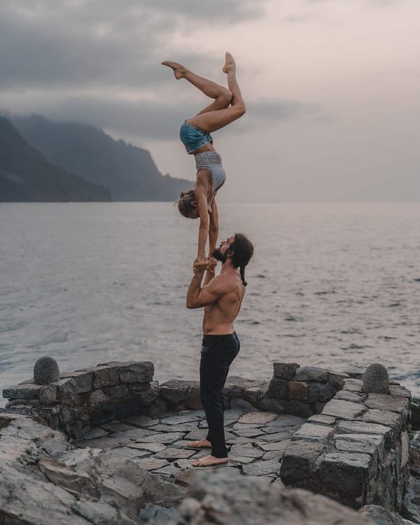 A man lifts a woman up with the strength of his arms in an acrobatic pose where she is holding herself upside down by her arm